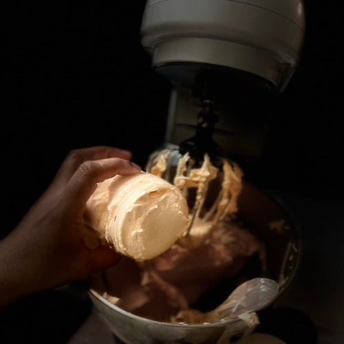 Hand holding a container of body butter over a bowl with a mixer in the background