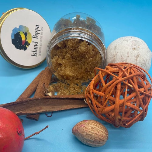 Open jar of pumpkin sugar scrub with a 'Island Peppa' label, cinnamon stick, nutmeg, red apple, and decorative ball on a blue background.